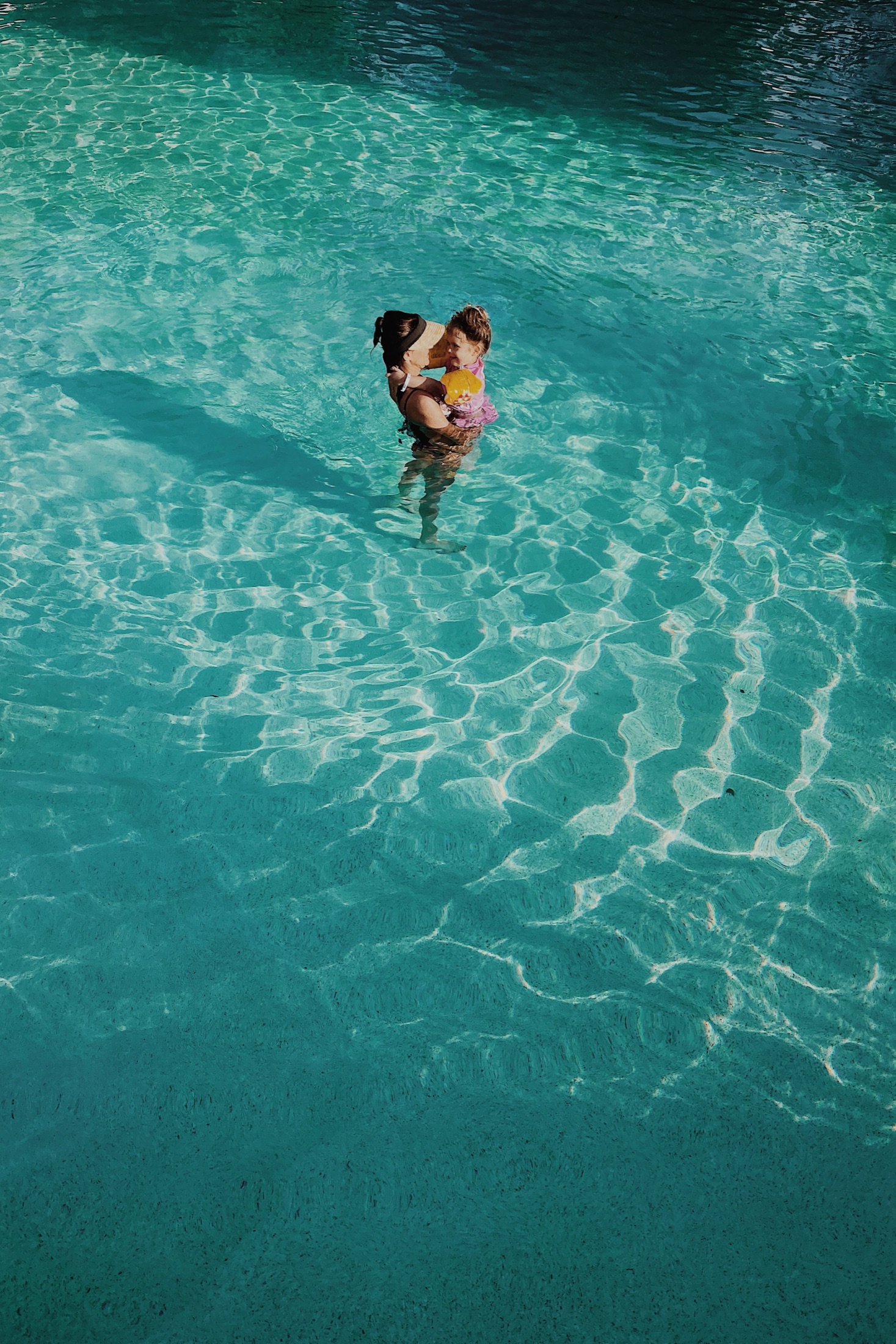 A mother holds her smiling child in sunlit turquoise pool water.