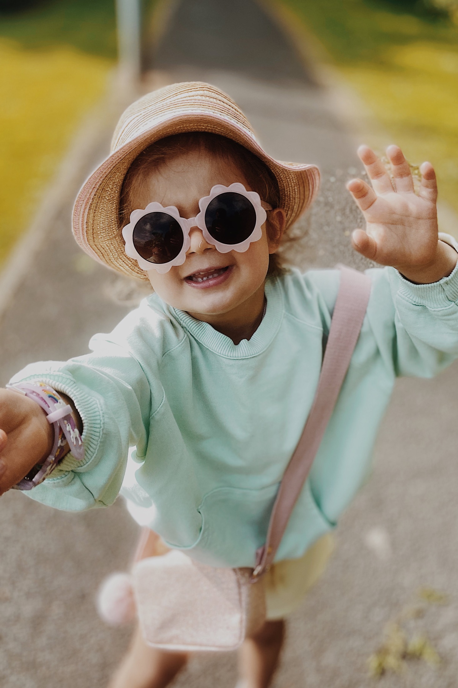 A child wearing flower-shaped sunglasses, a sunhat and a mint sweatshirt waves at the camera.